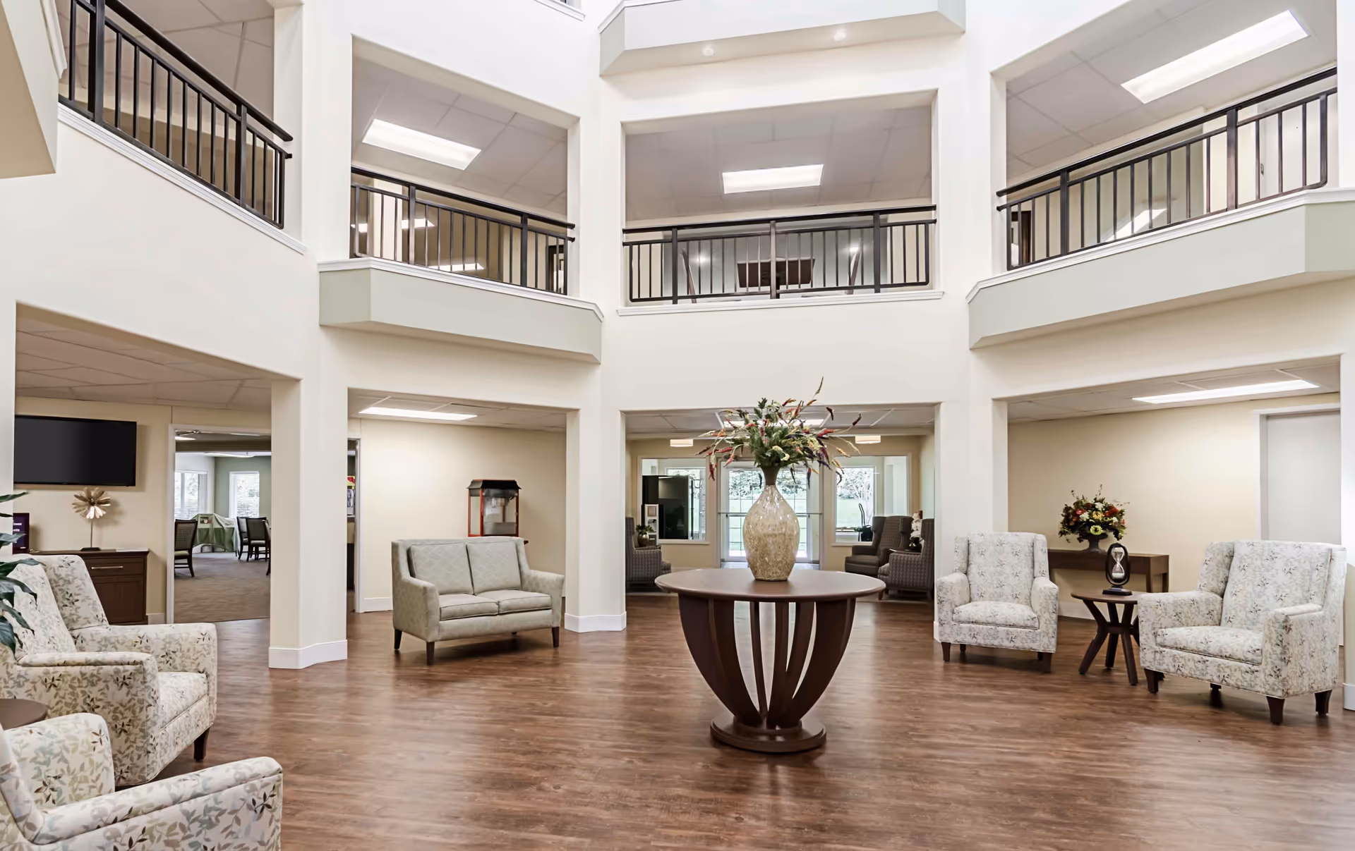 A spacious and well-lit senior living facility common area with a high ceiling and an open balcony on the second floor. The room features a round wooden table with a large decorative vase of flowers in the center. Surrounding the table are several upholstered armchairs and loveseats in light, neutral tones with floral patterns. The flooring is wood, and there are large windows and doorways leading to other rooms, including a dining area visible in the background.