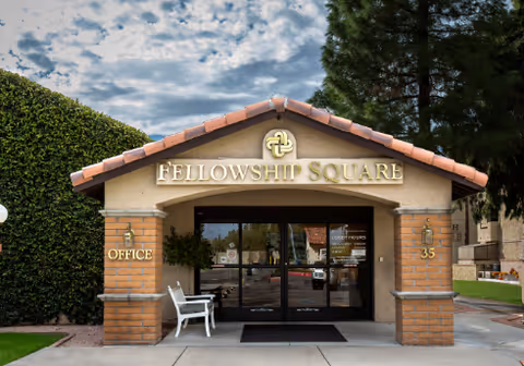 Entrance to Fellowship Square Historic Mesa facility with a tiled roof, brick pillars, glass double doors, a white bench on the left side, and the words 'FELLOWSHIP SQUARE' displayed above the entrance. The building number 35 is visible on the right pillar, and there is greenery including a tall hedge and trees around the entrance.