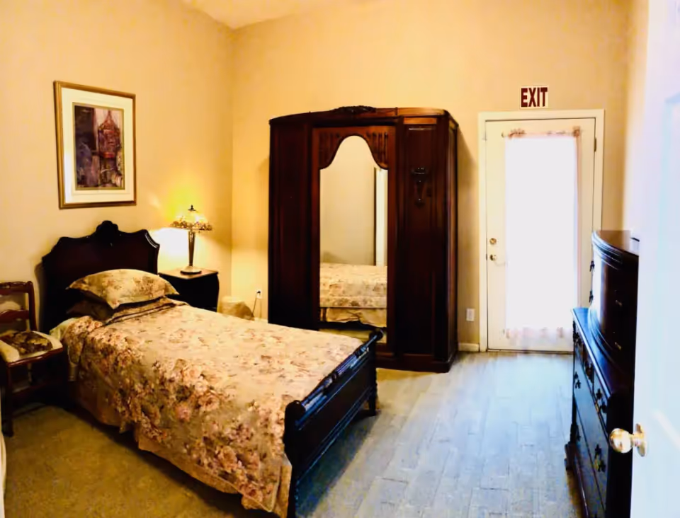 A cozy single-occupancy bedroom with a floral bedspread, wooden headboard and armoire with mirror, bedside lamp and an exit door.