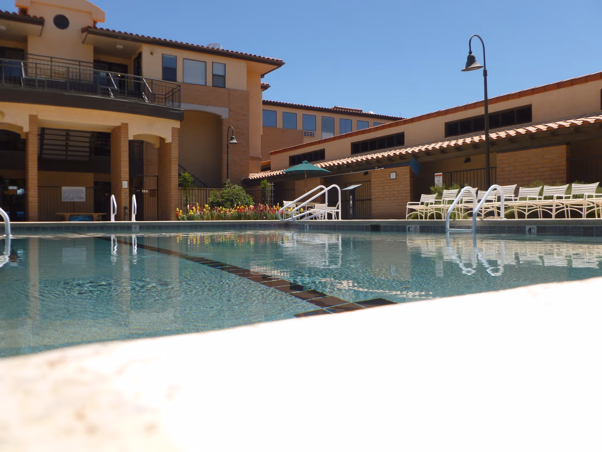 Outdoor swimming pool with clear water surrounded by a building with balconies and patio chairs under a clear blue sky.