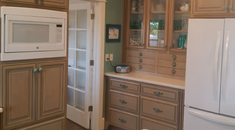 A kitchen area featuring wooden cabinets with turquoise knobs, a built-in microwave, a white refrigerator, and a countertop with a small radio. There is a glass-paneled door leading to another room.
