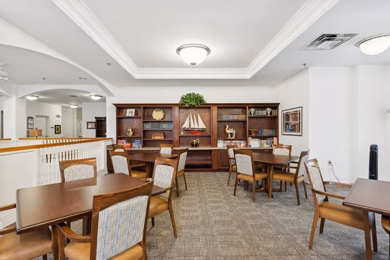 A well-lit common area with several wooden tables and chairs arranged on a carpeted floor. In the background, there is a large wooden bookshelf filled with books, decorative items including a model sailboat, and board games. The walls are white, and the ceiling has recessed lighting and crown molding.