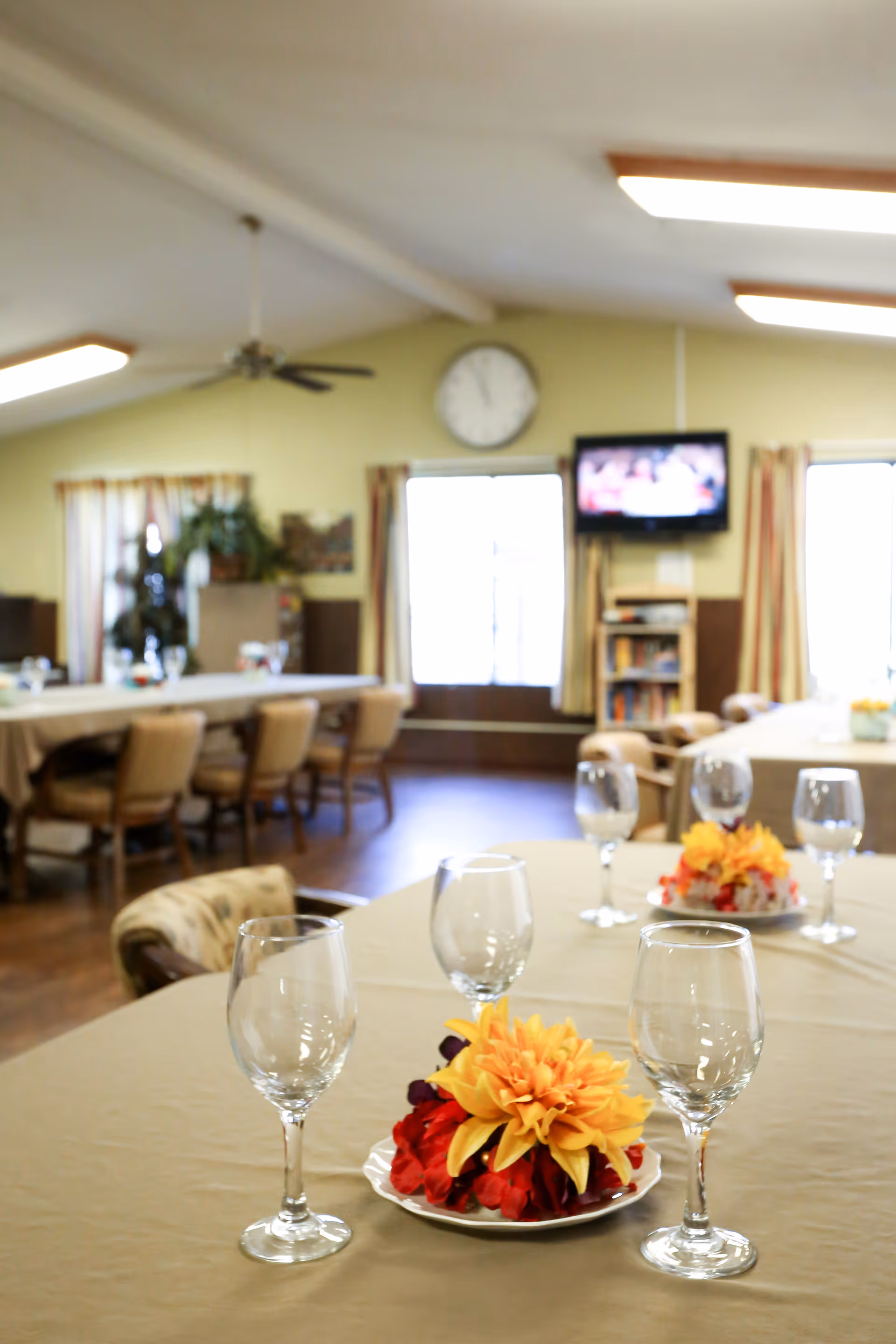 A dining room with tables covered in beige tablecloths, each set with empty wine glasses and a floral centerpiece featuring yellow and red flowers. The room has wooden chairs, large windows with curtains, a wall clock, a ceiling fan, and a wall-mounted television above a bookshelf.