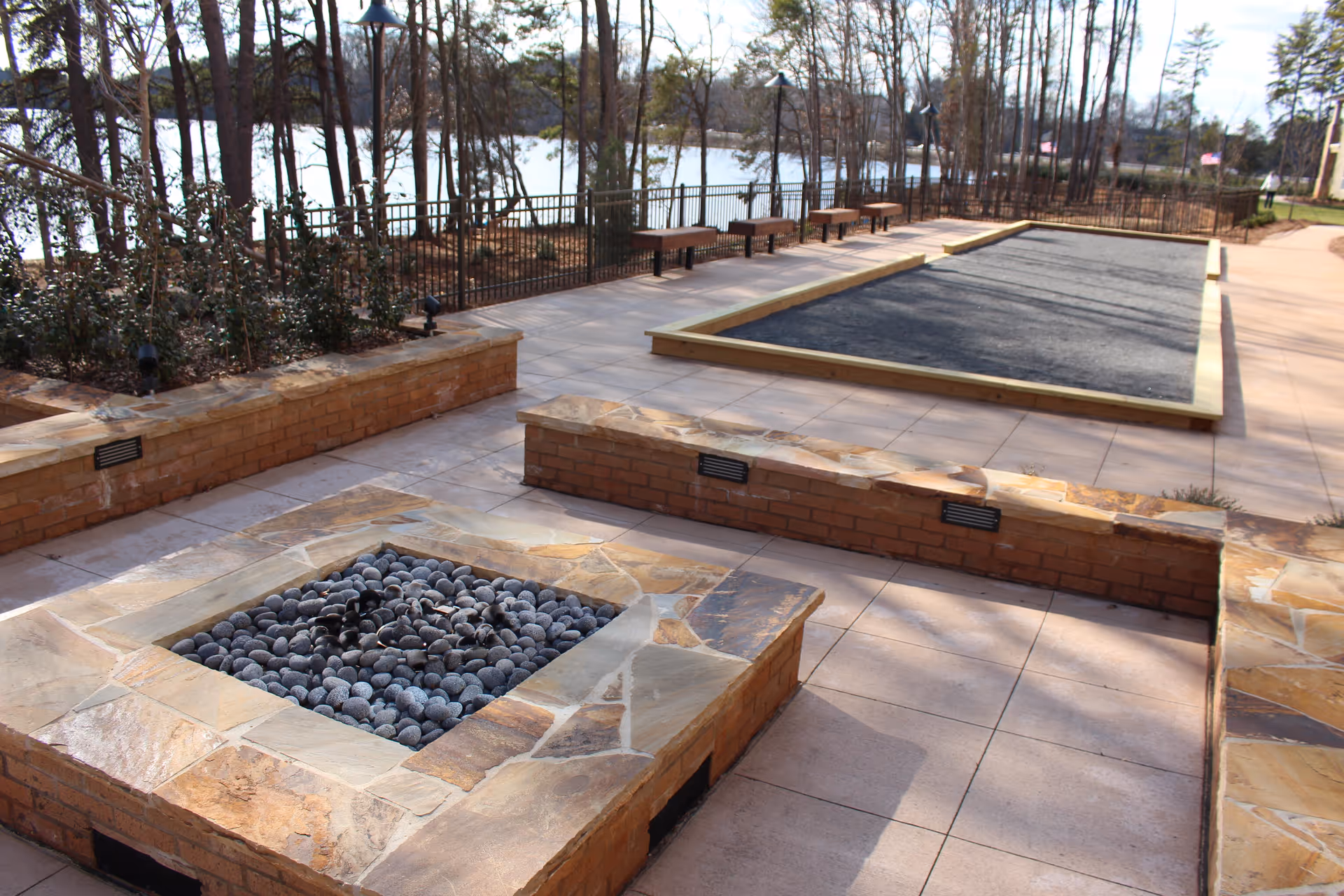 Outdoor patio area with a stone fire pit filled with black stones, surrounded by brick and stone seating walls. In the background, there is a bocce ball court and several benches along a fence with trees and a lake visible beyond.