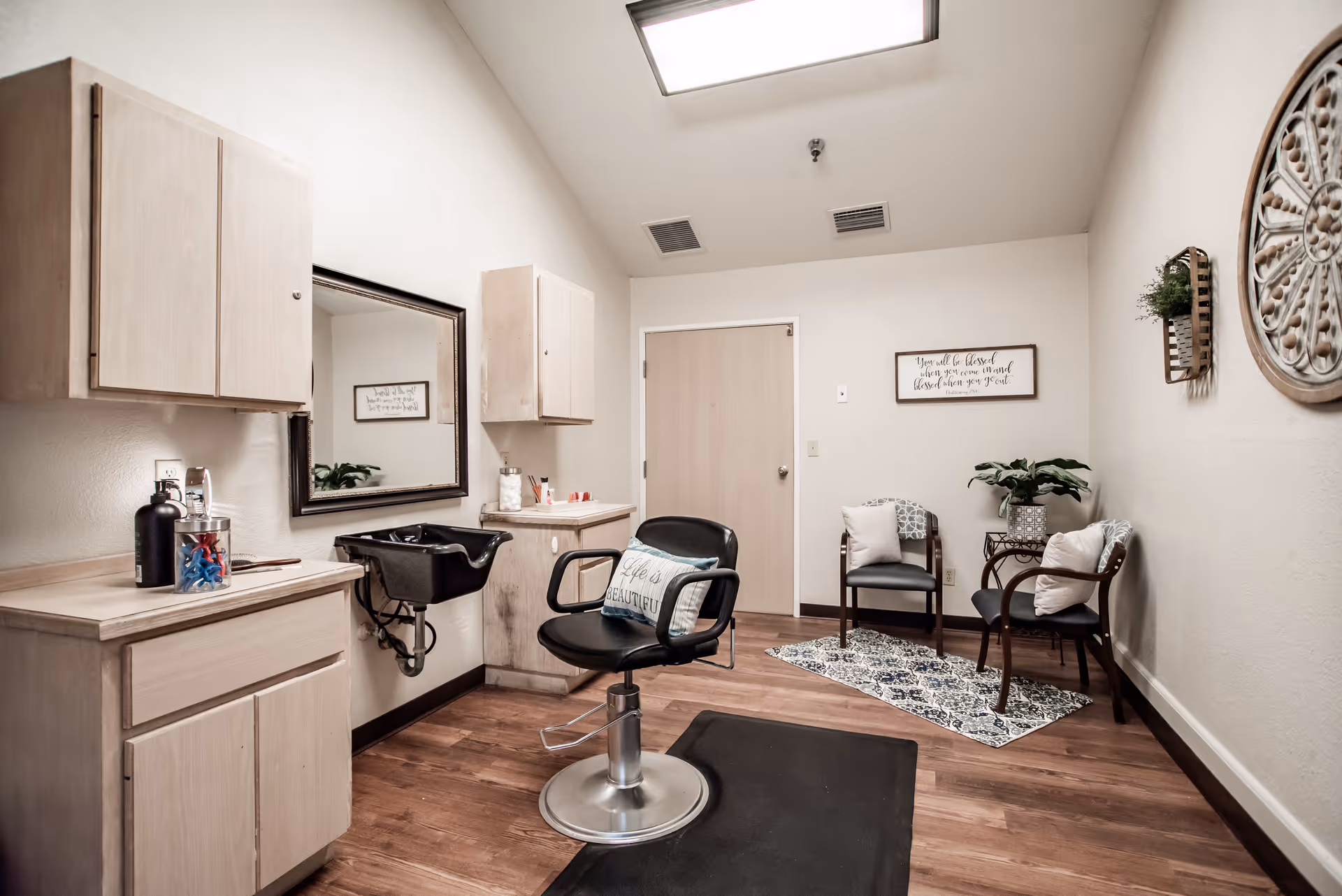 A small hair salon room with a black salon chair in front of a mirror and a black hair washing sink attached to the wall. There are light wood cabinets on the walls and a counter with hair products. The room has wooden flooring and a seating area with three chairs, cushions, a small table with a plant, and a decorative rug. The walls are decorated with framed quotes and a round ornamental wall piece.
