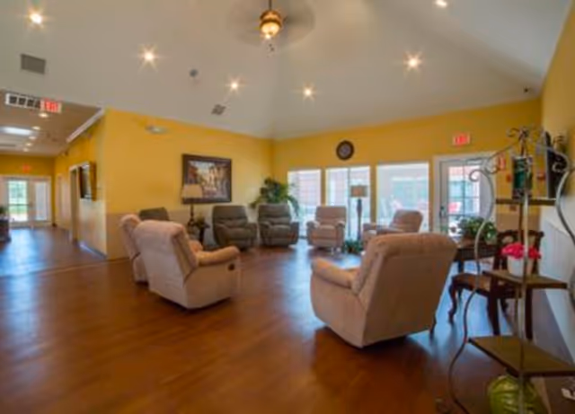 A spacious living room with yellow walls and wooden flooring, furnished with several cushioned armchairs arranged in a circle. The room has large windows letting in natural light, a clock on the wall, a framed picture, and some indoor plants. There is a hallway visible to the left and a door to the outside on the right.