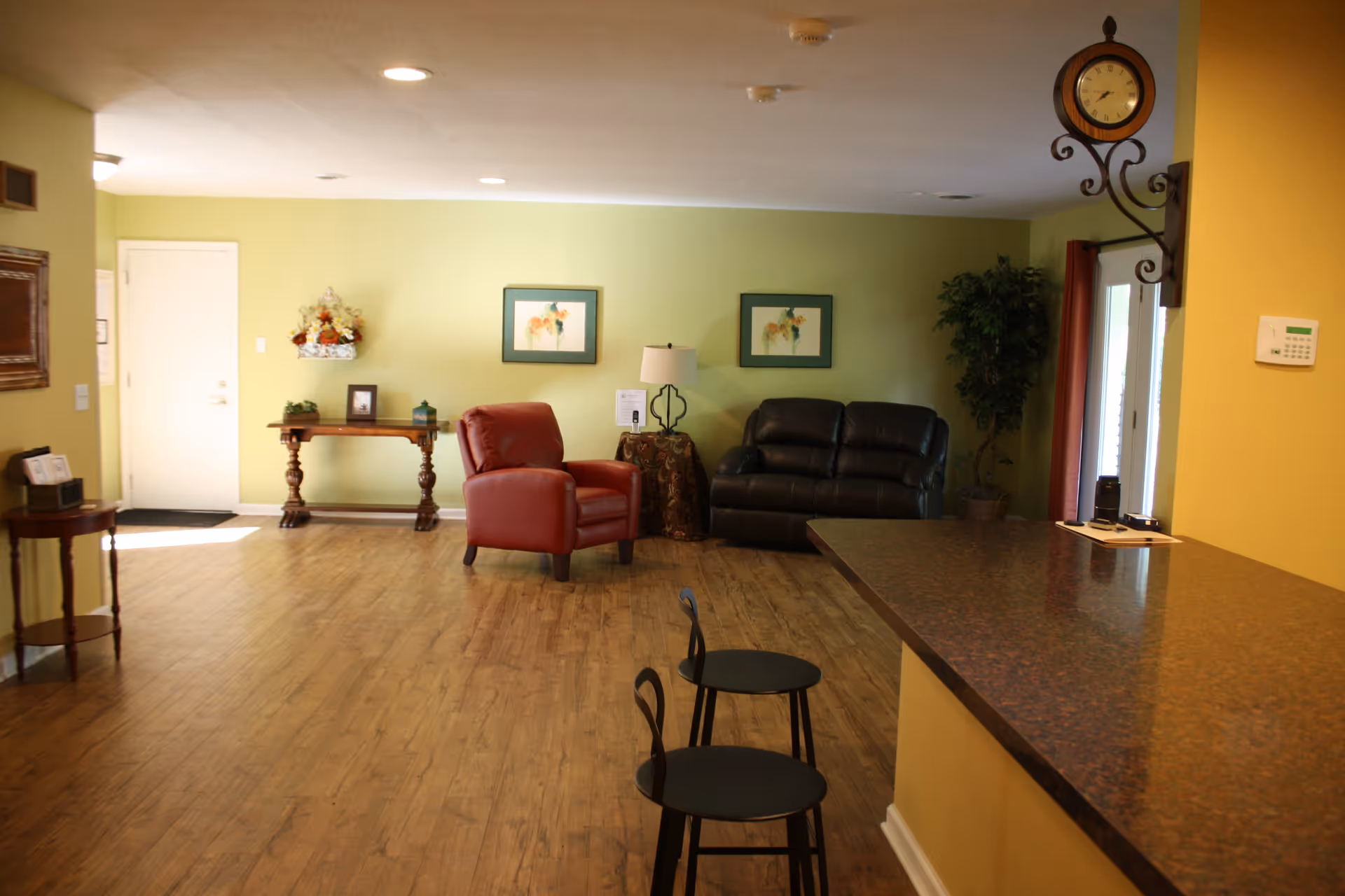 A cozy living room area with light green walls, wooden flooring, a red armchair, a black leather loveseat, a small round table with a lamp, two framed pictures on the wall, a wooden console table with decorative items, two black stools at a brown countertop, and a wall-mounted clock.