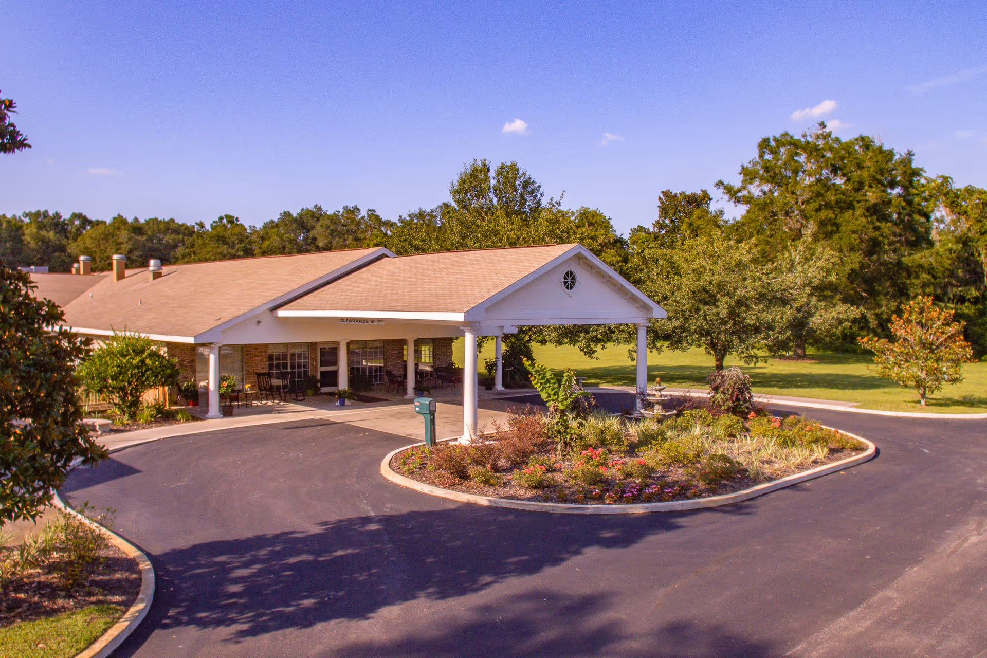 Front exterior view of Lafayette Nursing and Rehabilitation facility showing a covered entrance with white pillars, a circular driveway, landscaped garden with flowers and shrubs, and a green lawn with trees in the background under a clear blue sky.