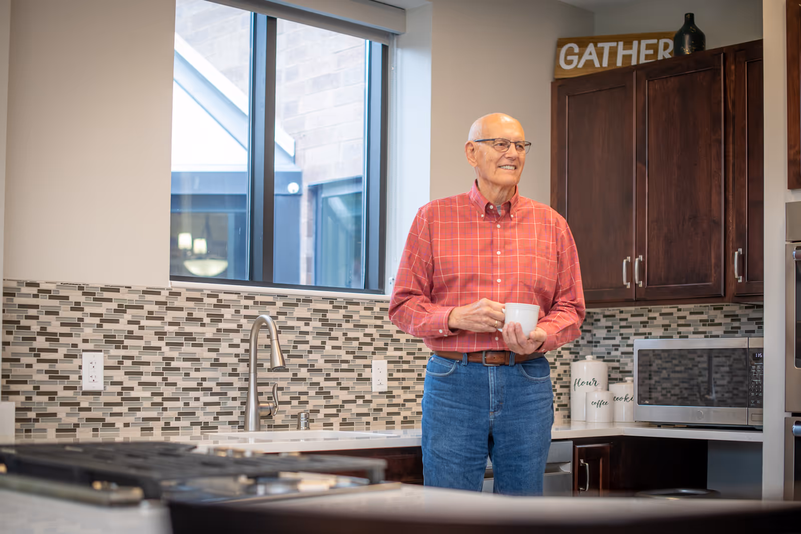 An older man holding a mug stands in a modern kitchen with a mosaic tile backsplash, dark cabinets, and a 'GATHER' sign.