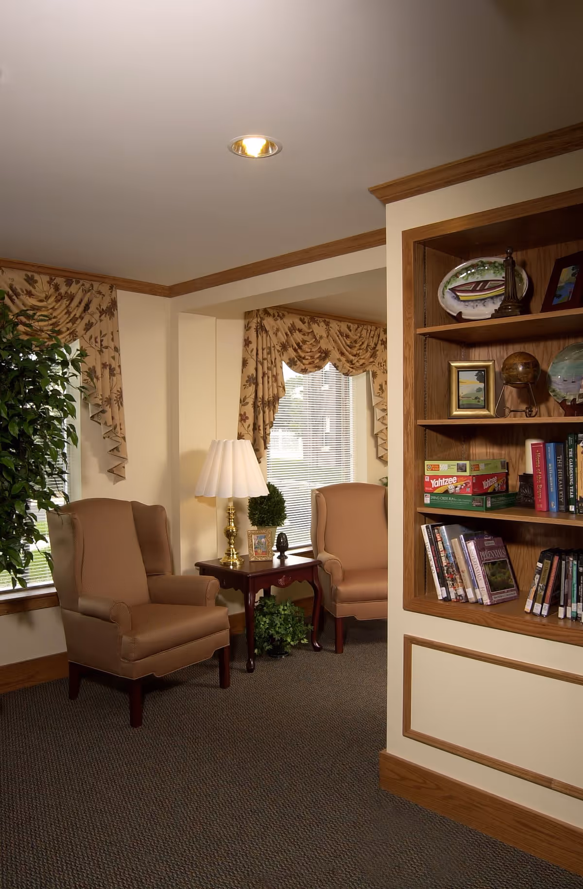 Cozy sitting area with two upholstered armchairs, a side table and lamp by a window with patterned valances, and a built-in bookshelf.