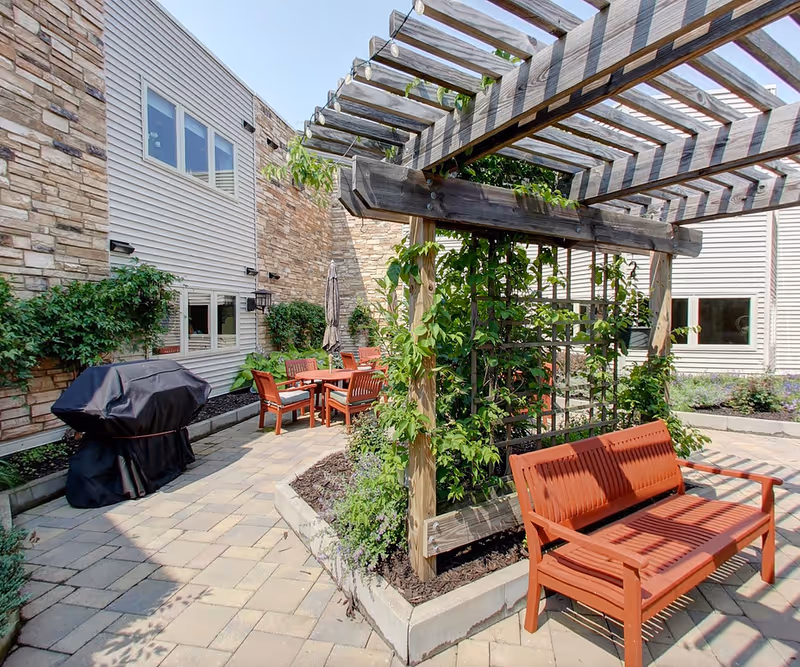 Outdoor patio area at Prairie Hills at Cedar Rapids featuring a wooden pergola with climbing plants, a red wooden bench, a covered grill, and a table with chairs surrounded by stone and siding building walls.