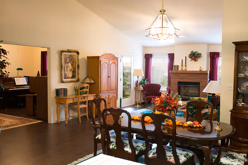 Bright open living and dining area with a wooden dining table and chairs, a fireplace, armchairs, and a piano visible in an adjacent room.