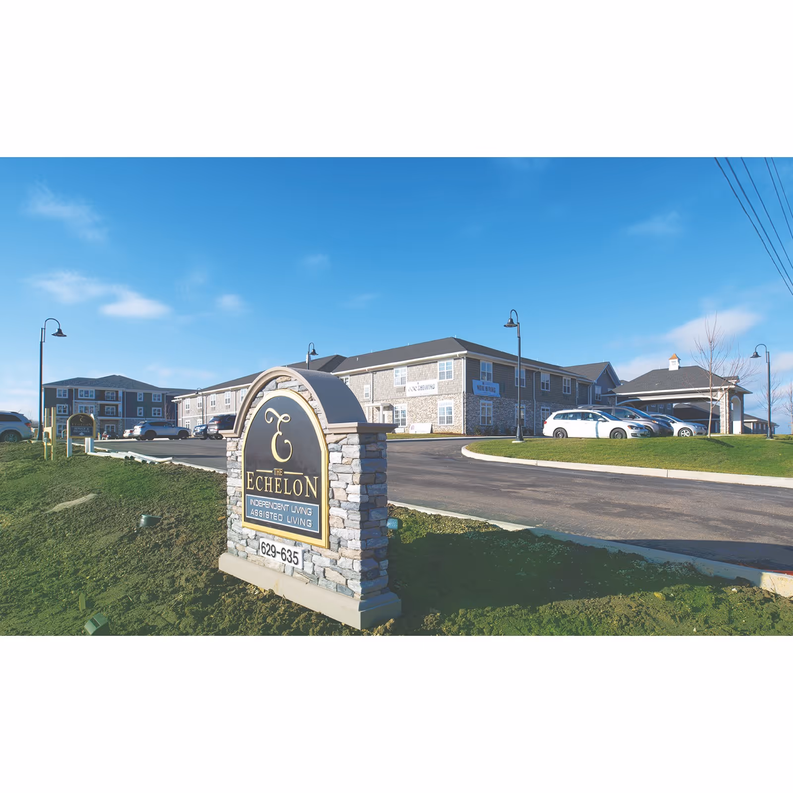 Exterior view of The Echelon of Medina senior living facility on a clear day, showing a stone sign with the facility's name and services offered, a paved driveway, parked cars, and a two-story building in the background.