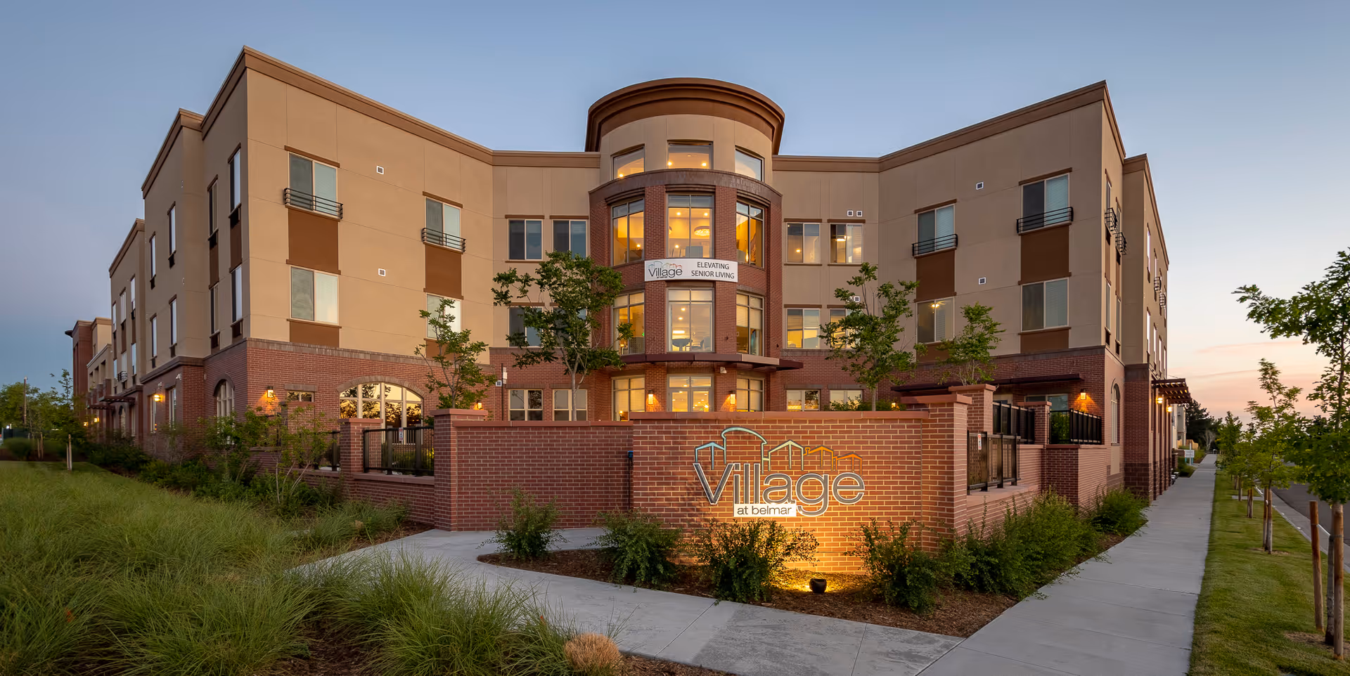 Exterior view of a modern three-story senior living facility named Village at Belmar during dusk. The building features a combination of brick and beige siding with multiple windows and a prominent rounded central section with large windows. There is a brick wall in front with the illuminated sign 'Village at Belmar' and landscaped greenery along the sidewalk.