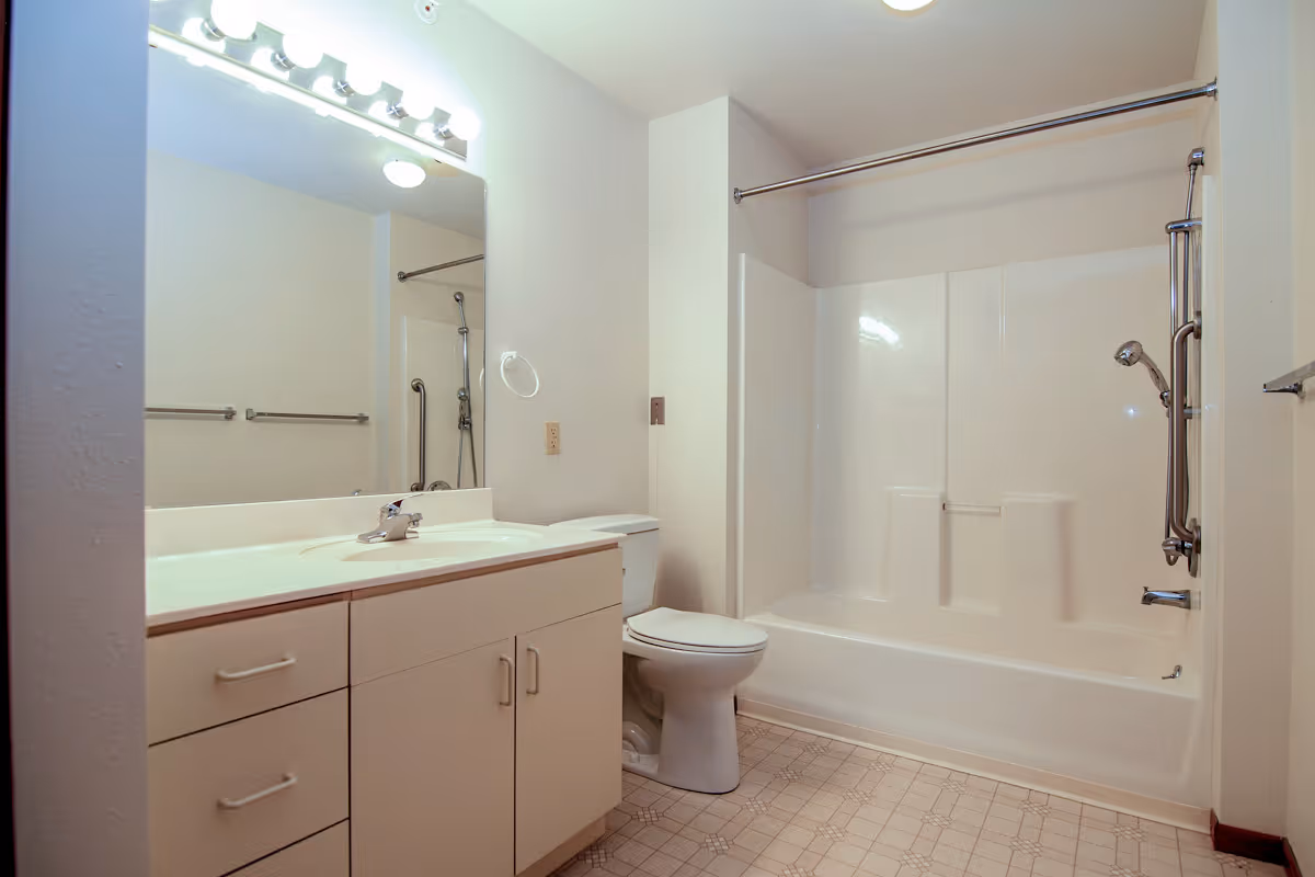 A clean bathroom featuring a white vanity with a sink and mirror above it, a toilet, and a bathtub with a showerhead and grab bars. The floor has a patterned tile design and the walls are painted light beige.