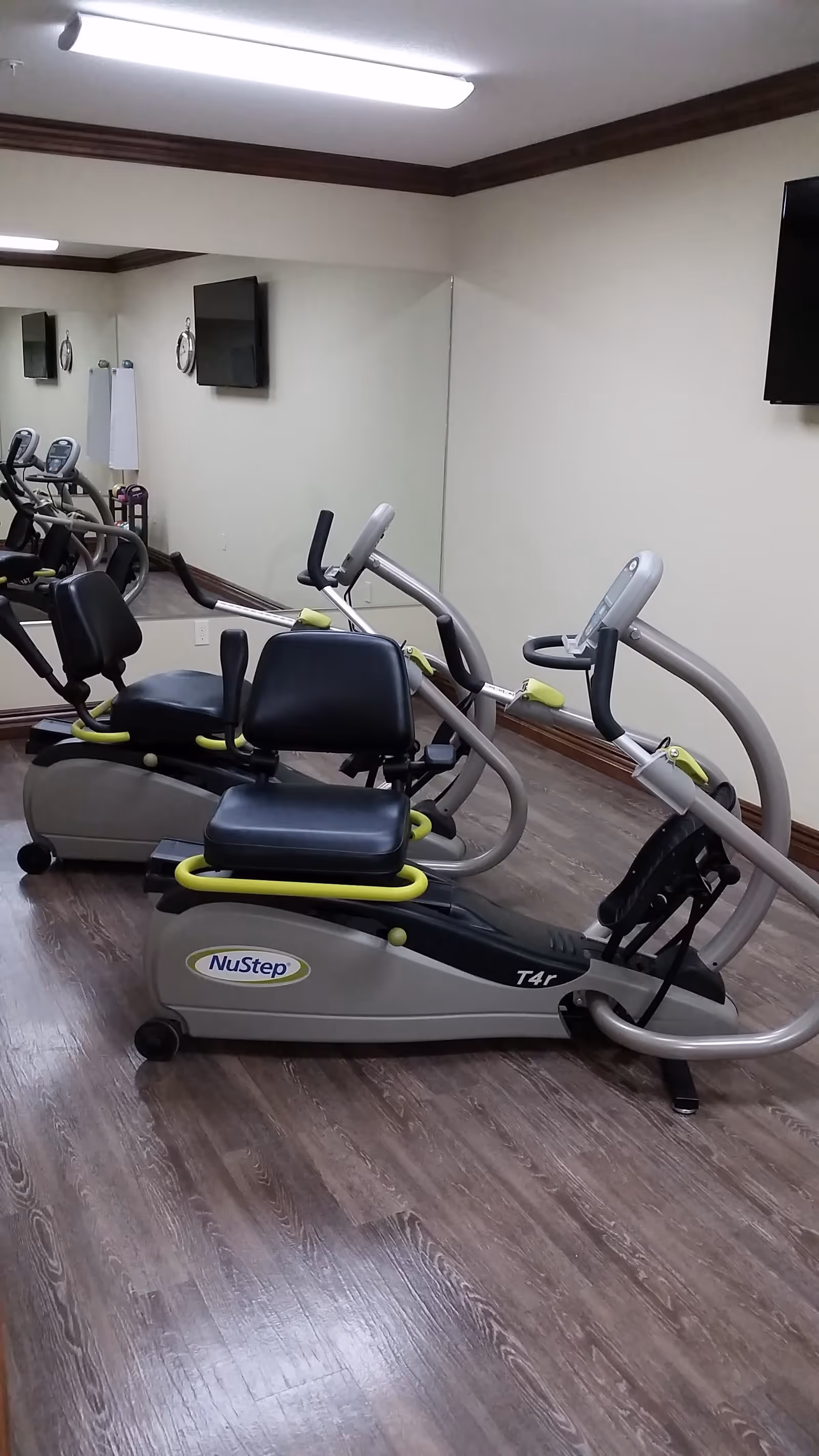 A small exercise room with two NuStep recumbent cross trainers positioned on a wood floor. The room has beige walls with dark wood trim, a large wall mirror, and a wall-mounted TV.