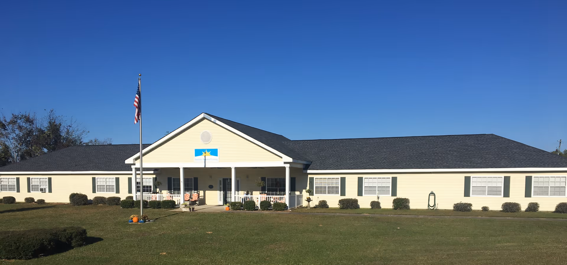 Exterior view of a single-story assisted living facility building with light yellow siding and a dark roof. The building has multiple windows with green shutters and a covered entrance porch with white columns. An American flag is flying on a flagpole in front of the building, and there is a well-maintained lawn with some small bushes and a few pumpkins near the entrance. The sky is clear and blue.