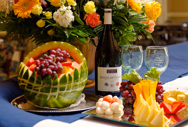A decorative buffet arrangement with a carved watermelon filled with fruit, a bottle of wine, two wine glasses, and a platter of cheeses and grapes beside a vase of flowers.