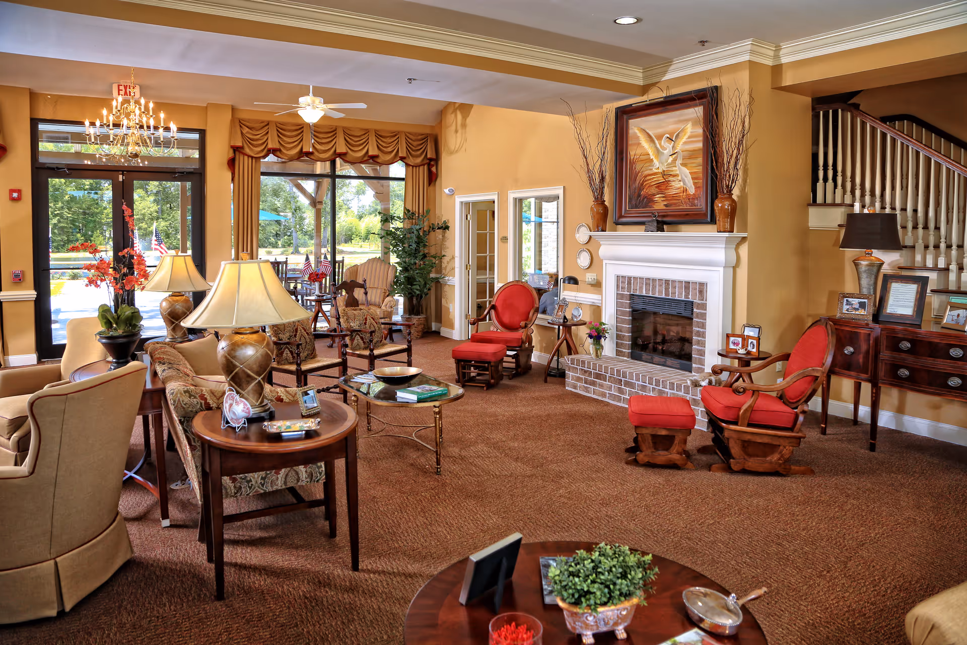 A cozy senior living facility common area with warm beige walls and carpet. The room features multiple comfortable chairs and sofas arranged around a brick fireplace with a painting of two white birds above it. There are several side tables with lamps, plants, and framed photos. Large windows and glass doors let in natural light and offer a view of the outdoor greenery. A staircase is visible in the background.
