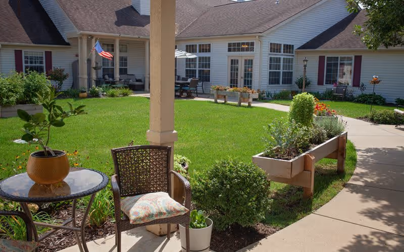 Outdoor courtyard area of a senior living facility with a green lawn, raised garden beds, patio furniture including a glass table with a potted plant and a chair with a cushion, surrounded by a white building with windows and a walkway.