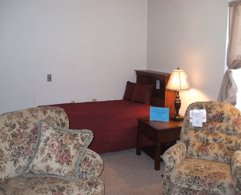 Small bedroom with a single bed covered in a burgundy bedspread, a nightstand with a lamp, and two floral upholstered chairs.