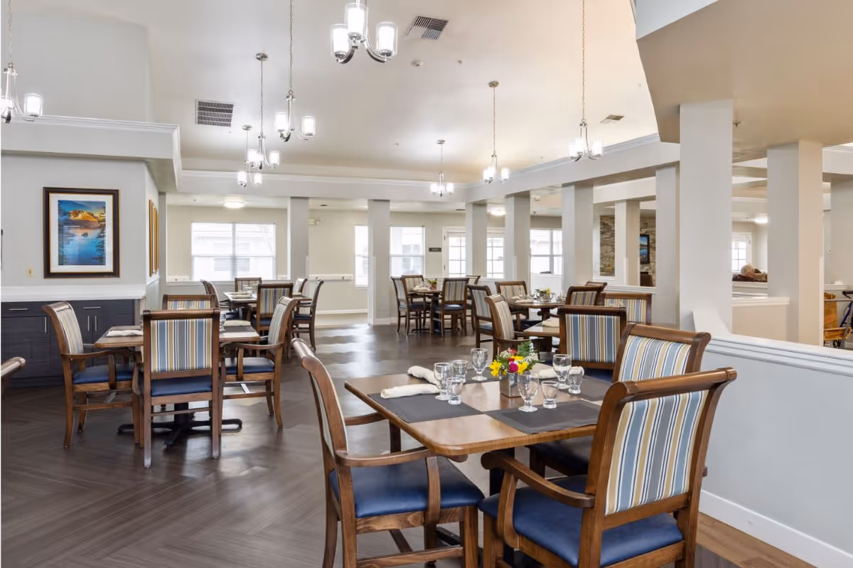 A bright and spacious dining room with multiple wooden tables and chairs. Each table is set with placemats, glassware, and napkins, and some have small flower arrangements. The room features large windows allowing natural light, modern hanging light fixtures, and framed artwork on the walls.
