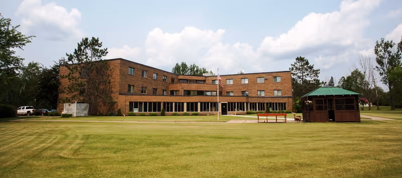 A three-story brick building with multiple windows, surrounded by a large grassy lawn. There is an American flag on a flagpole in front of the building, a wooden gazebo with a green roof to the right, and some trees in the background under a partly cloudy sky.