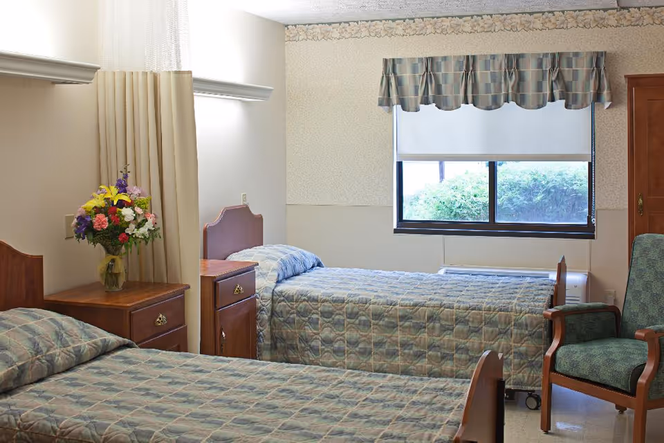 A simple bedroom in a senior living facility with two single beds covered in matching patterned bedspreads. Between the beds is a wooden nightstand with a vase of colorful flowers. A window with a patterned valance lets in natural light, and a green upholstered armchair is positioned near the window.