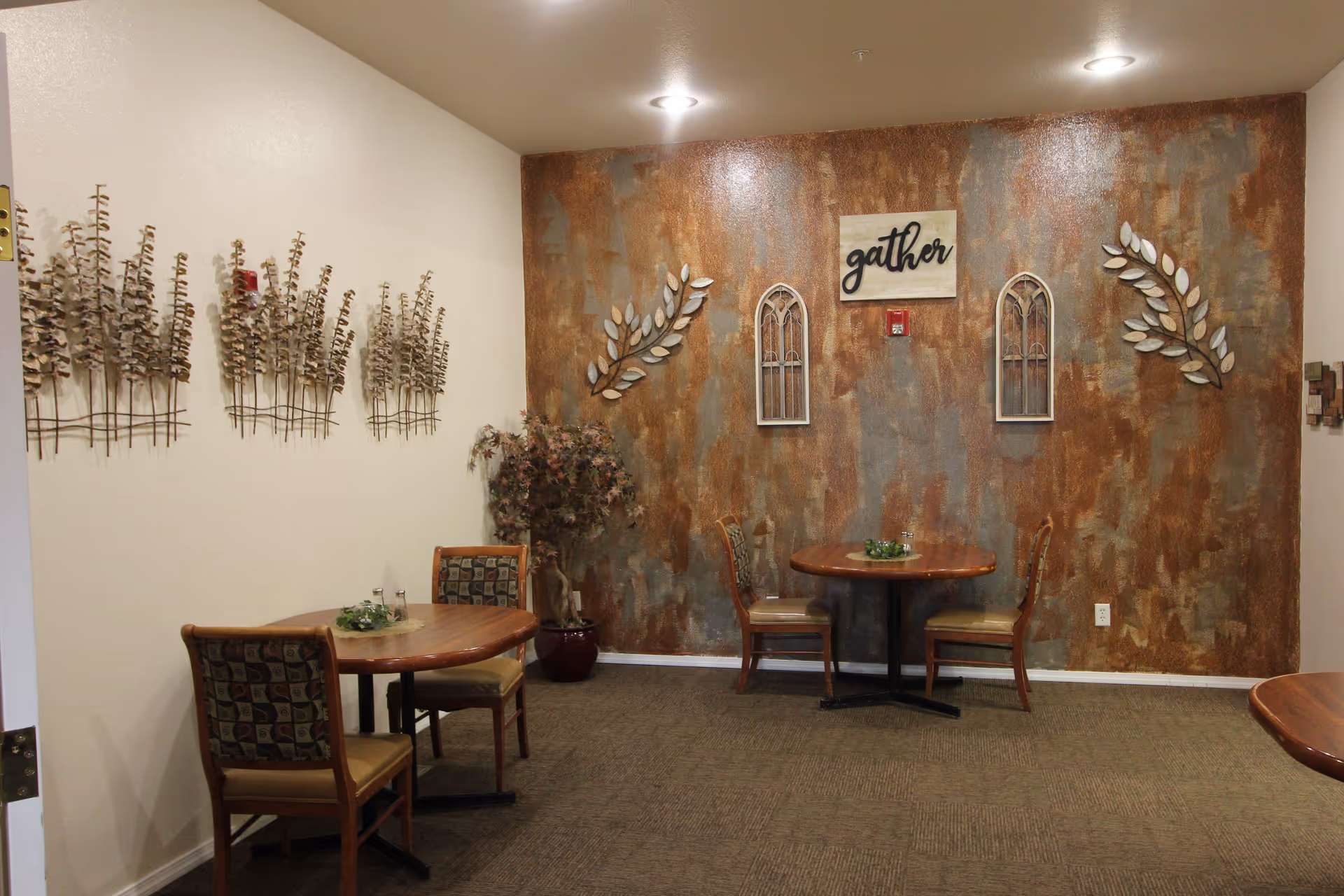 A cozy dining area with two round wooden tables, each accompanied by two chairs with patterned upholstery. The back wall features a textured design with decorative wall art including two arched window frames, leaf motifs, and a sign that says 'gather'. There is a potted plant in the corner and soft overhead lighting.