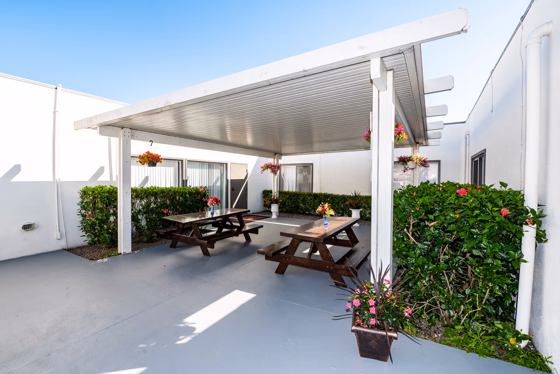 Covered outdoor courtyard with wooden picnic tables, hanging flower baskets, and surrounding hedges.