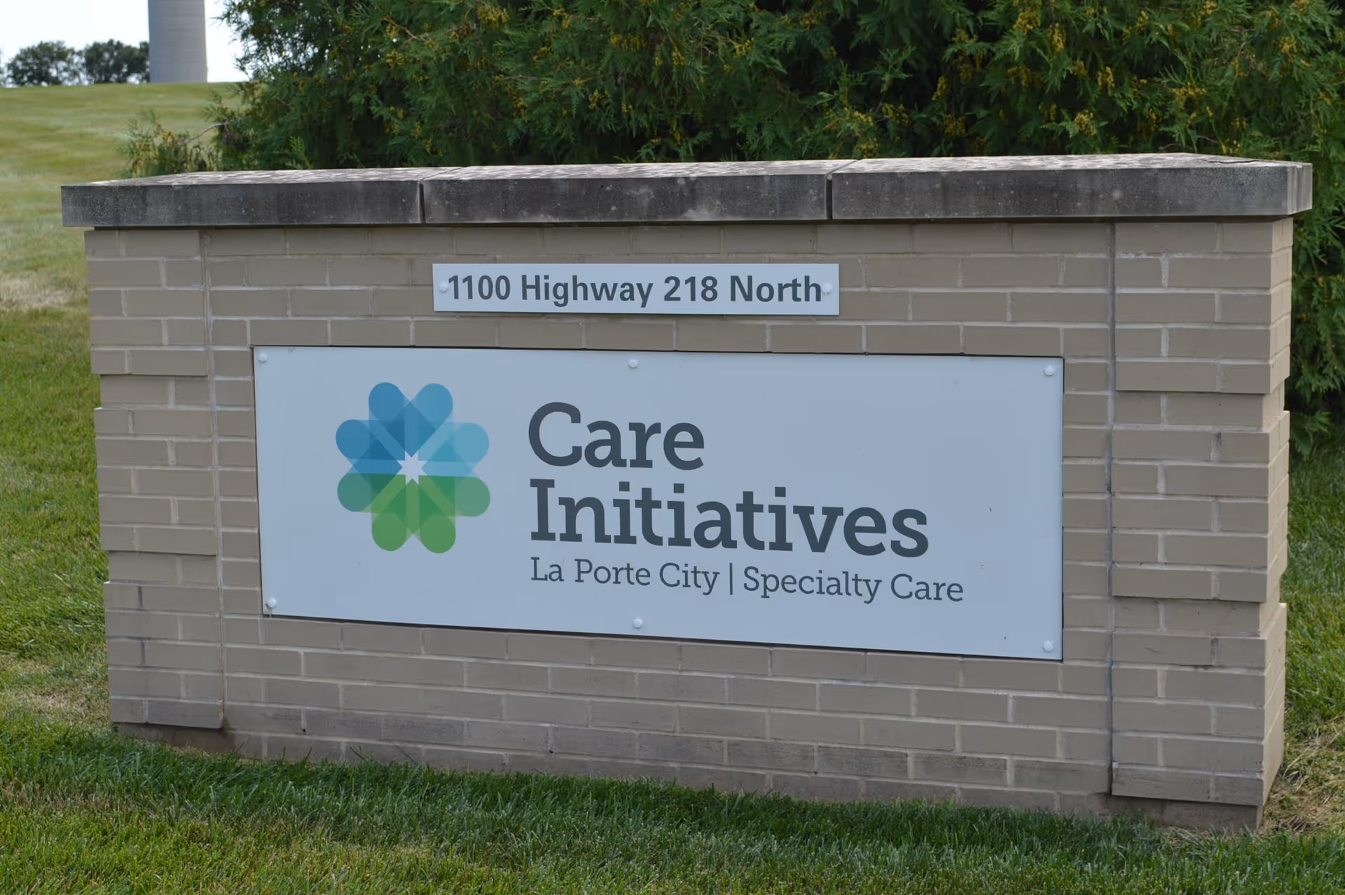 A brick signpost outdoors with a white sign displaying the logo and text 'Care Initiatives La Porte City | Specialty Care' and an address plaque above it reading '1100 Highway 218 North'.