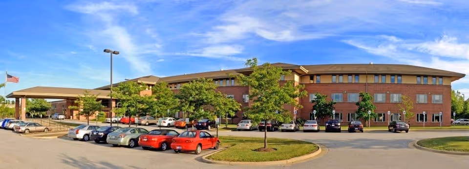 Front view of a large red-brick nursing and rehabilitation building with a covered entrance, parked cars, trees, and an American flag under a blue sky.