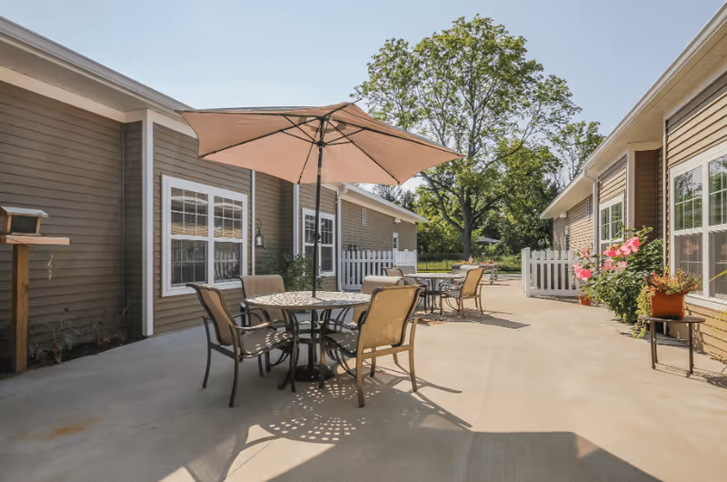 Outdoor patio area between two buildings with round tables, chairs, and large umbrellas. There are potted plants and flowers along the building walls, and a large tree is visible in the background under a clear sky.