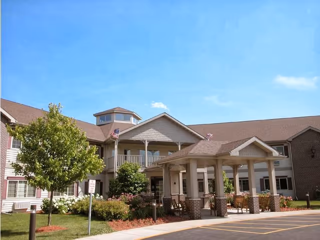 Front entrance of a senior living community building with a covered portico, outdoor seating, American flags, and landscaped grounds under a clear blue sky.