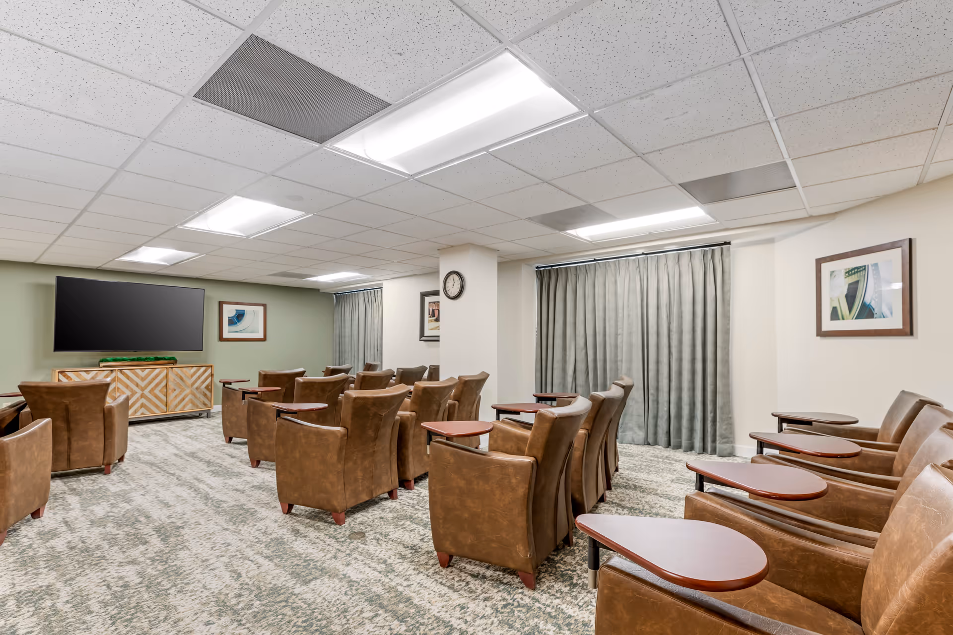 A small theater or media room with several rows of brown leather chairs, each equipped with a small wooden side table. The room has a carpeted floor, a drop ceiling with fluorescent lighting, a large flat-screen TV mounted on a green accent wall, and framed artwork on the walls. There are two windows covered with gray curtains and a clock mounted on a white column in the center of the room.