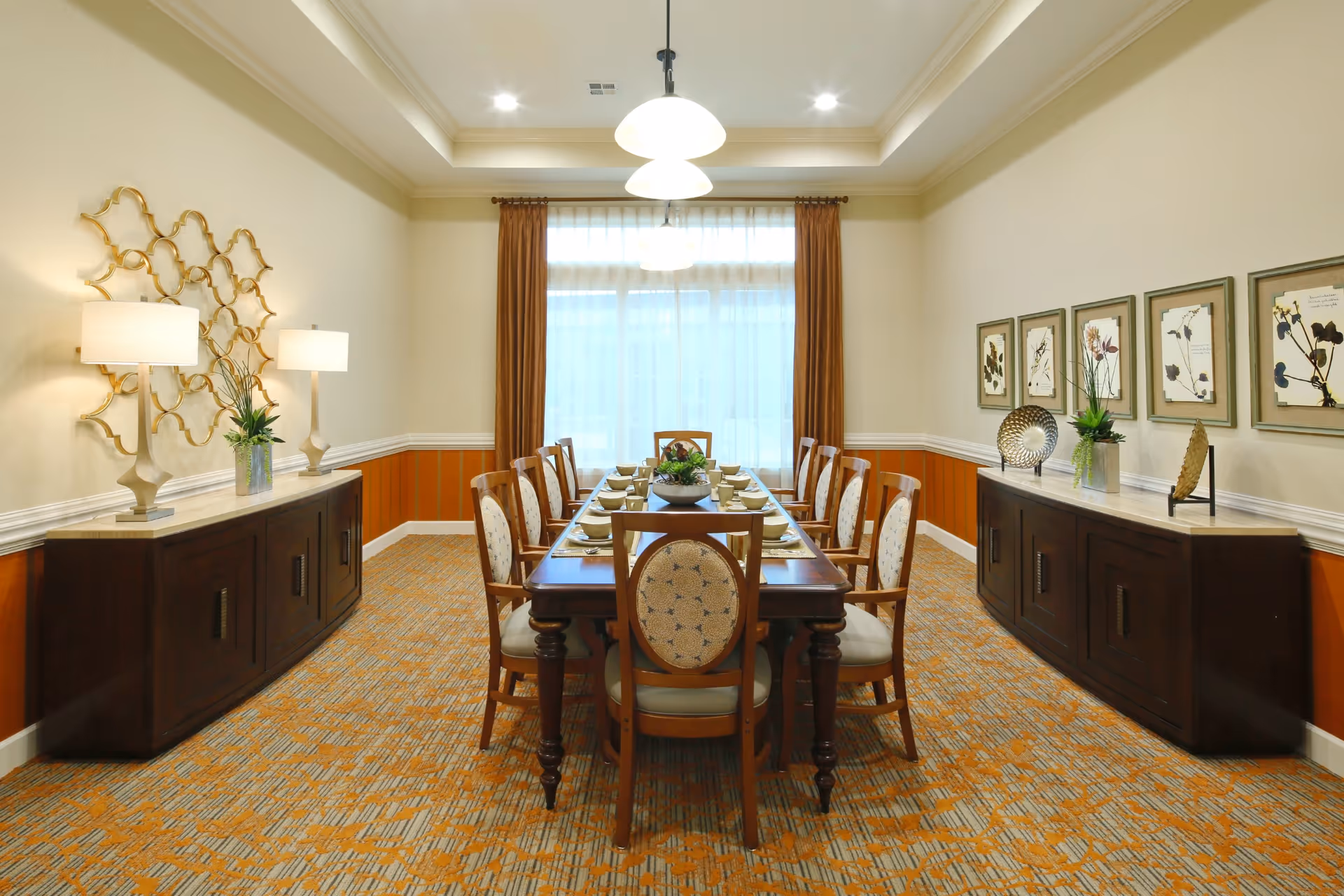 A formal dining room with a long wooden dining table set for ten people with matching chairs. The room features patterned carpet, two sideboards with decorative lamps, plants, and artwork on the walls. Large windows with sheer curtains and brown drapes allow natural light to fill the space.