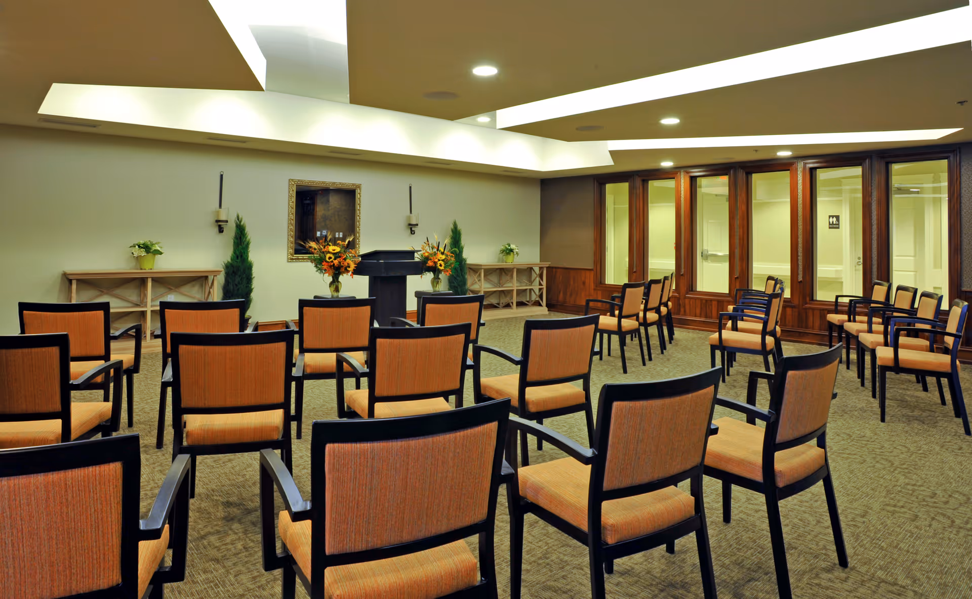 Spacious interior meeting room with rows of orange-upholstered chairs facing a lectern and floral arrangements.