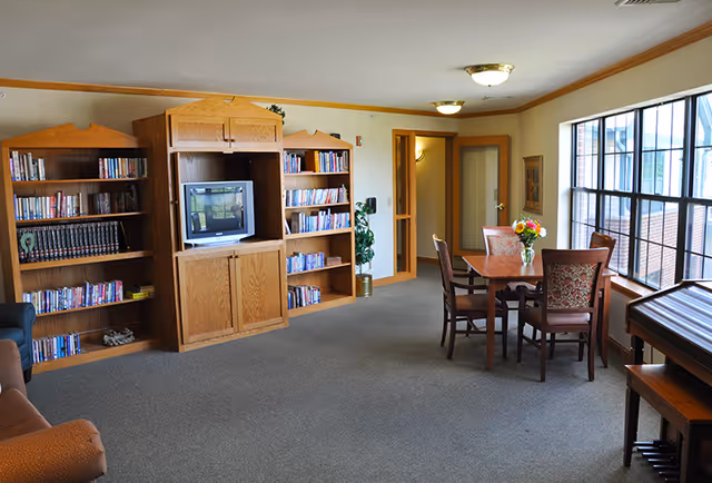 Well-lit common room with bookshelves and a TV on the left and a table with chairs beside large windows on the right.
