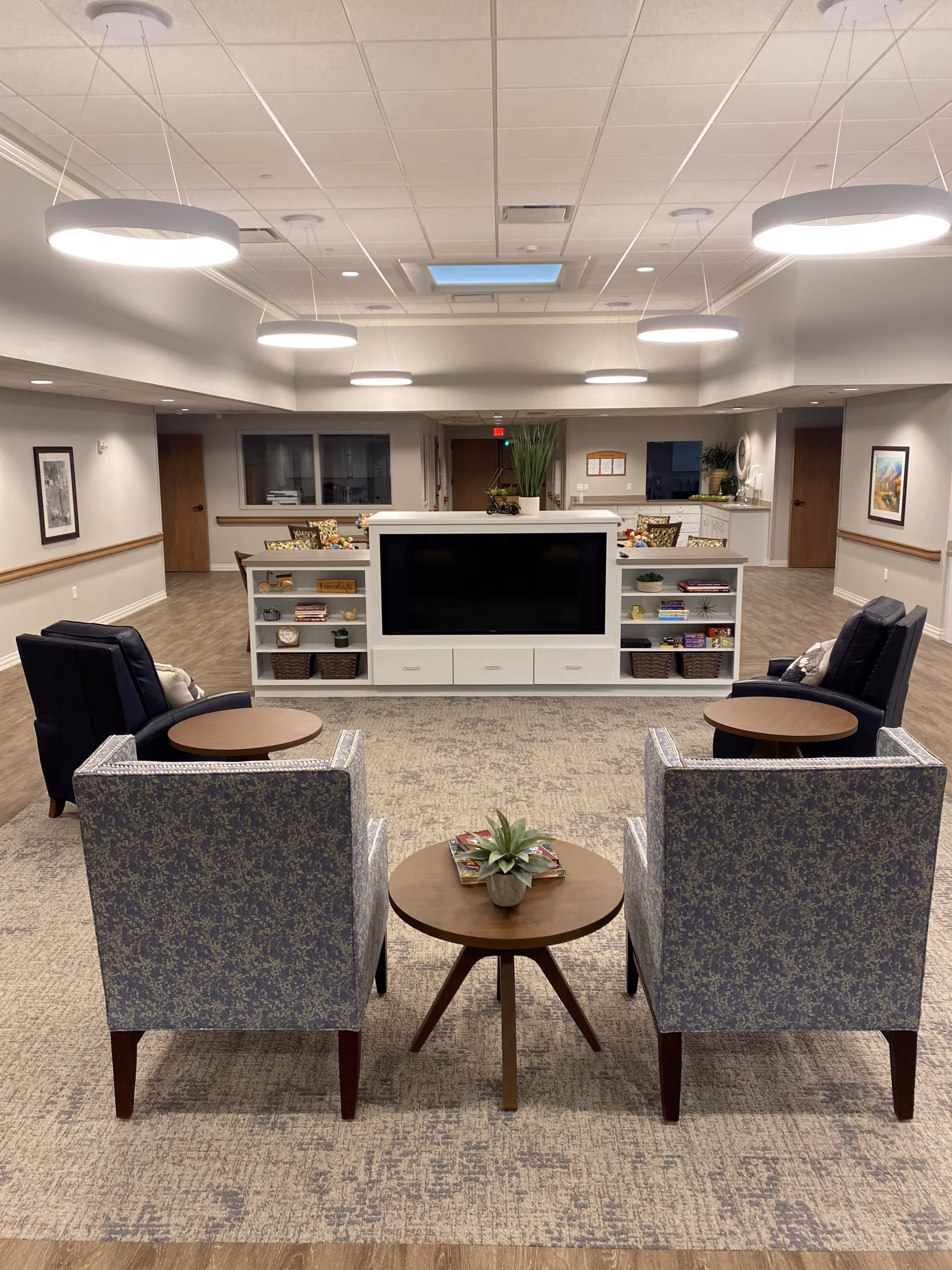 A spacious and well-lit living room area with four armchairs arranged around two small round wooden tables. In the center is a white entertainment unit with a large flat-screen TV and shelves holding books and decorative items. The room has a light-colored carpet and wooden flooring, with modern circular ceiling lights and a skylight above. Doors and framed artwork are visible on the walls in the background.