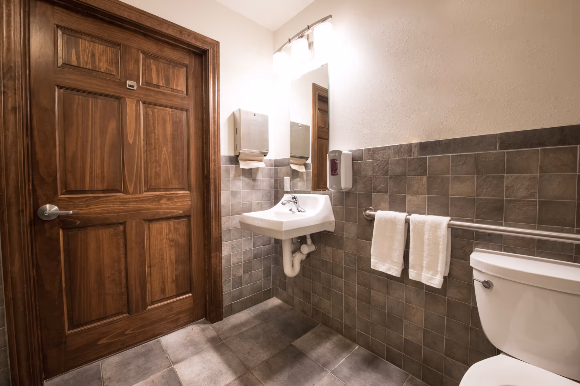 Accessible restroom with a wooden door, wall-mounted sink and mirror, toilet, grab bar, towel, and tiled walls and floor.