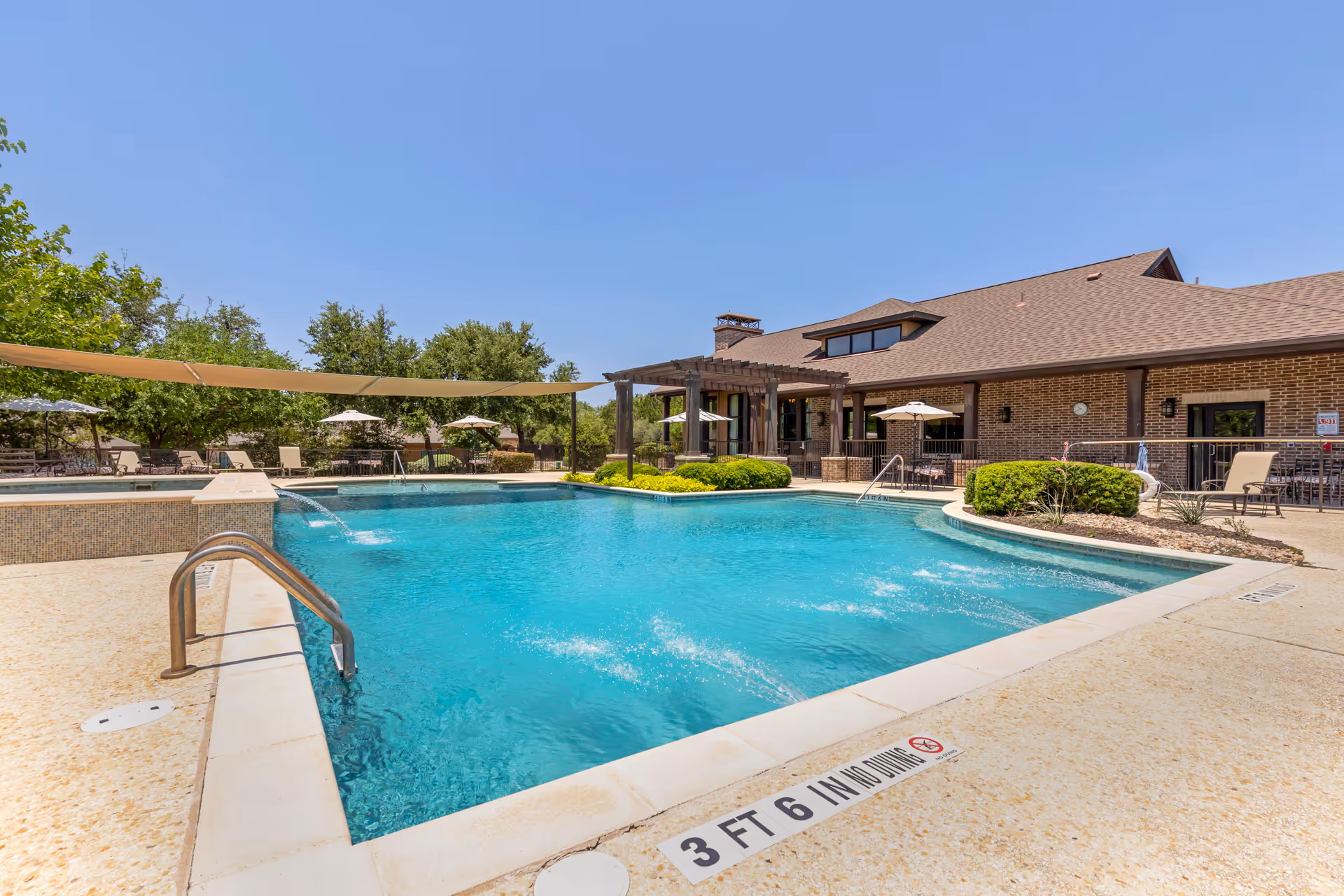 Outdoor swimming pool in front of a brick clubhouse with lounge chairs and umbrellas under a clear blue sky.