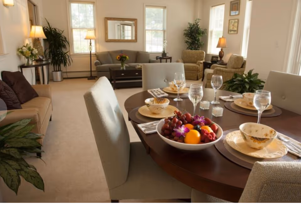 A cozy living and dining area with a round wooden dining table set for four, featuring plates, bowls, glasses, and a centerpiece bowl of fresh fruit and flowers. The background shows a living room with sofas, armchairs, lamps, plants, and windows letting in natural light.