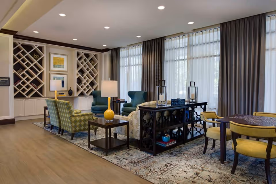 Bright assisted-living community lounge with armchairs, side tables, built-in shelving, and large curtained windows.