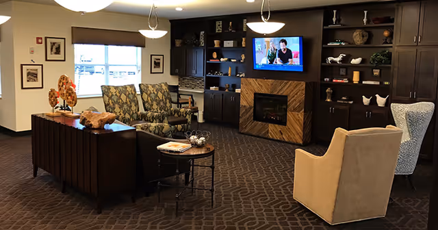 A cozy living room area with patterned carpet, several upholstered chairs, a dark wood cabinet with decorative items, a wall-mounted television above a modern fireplace, and a window with blinds letting in natural light.
