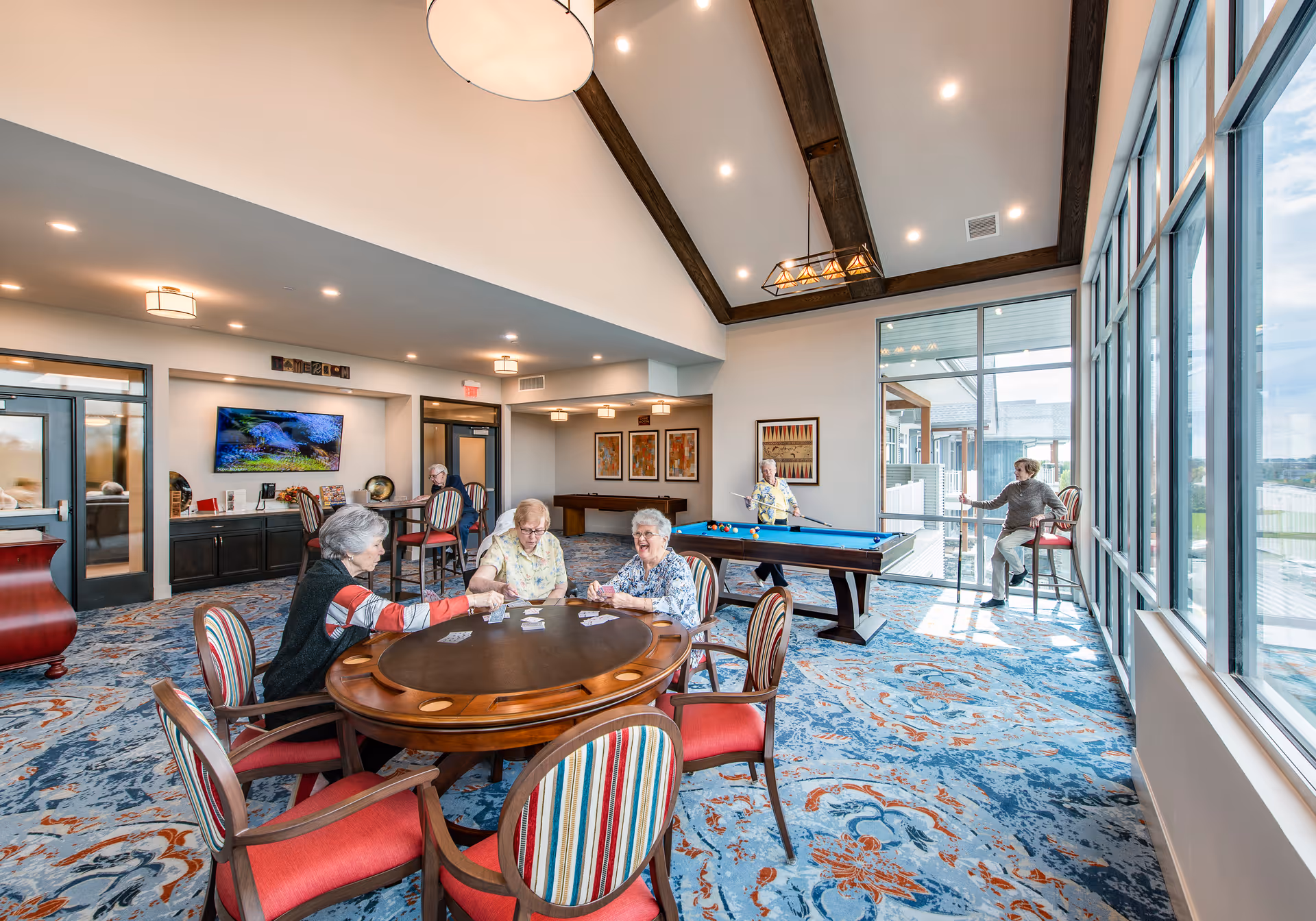 A bright and spacious senior living community room with large windows letting in natural light. Several elderly people are engaged in activities: three women are playing cards at a round wooden table with striped and red cushioned chairs, one woman is playing pool, and another woman is sitting on a high chair near the window. The room features a patterned blue and orange carpet, vaulted ceiling with wooden beams, and modern lighting fixtures.