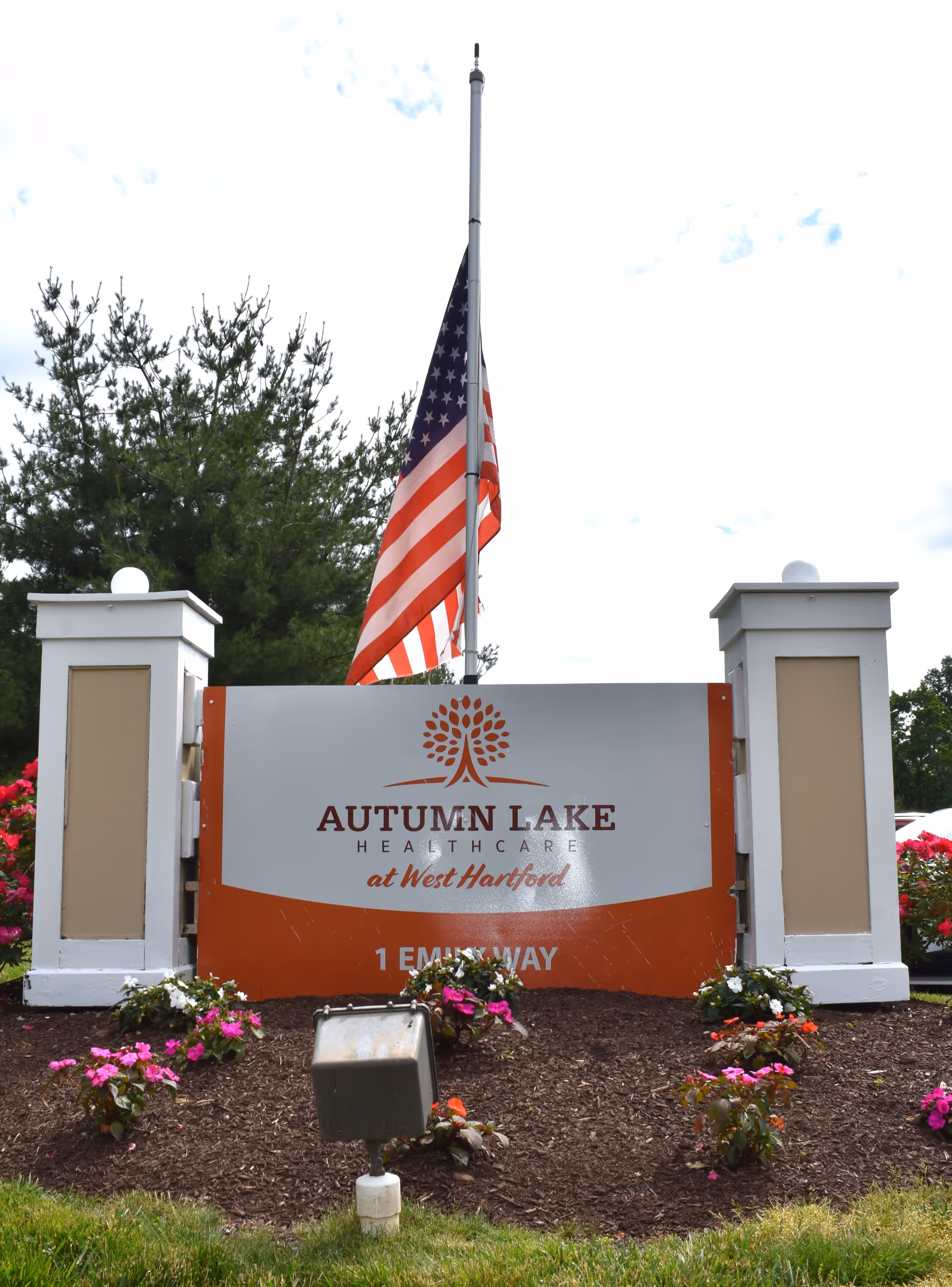 Outdoor sign for Autumn Lake Healthcare at West Hartford with an American flag flying at half-mast behind it, surrounded by flower beds and greenery.