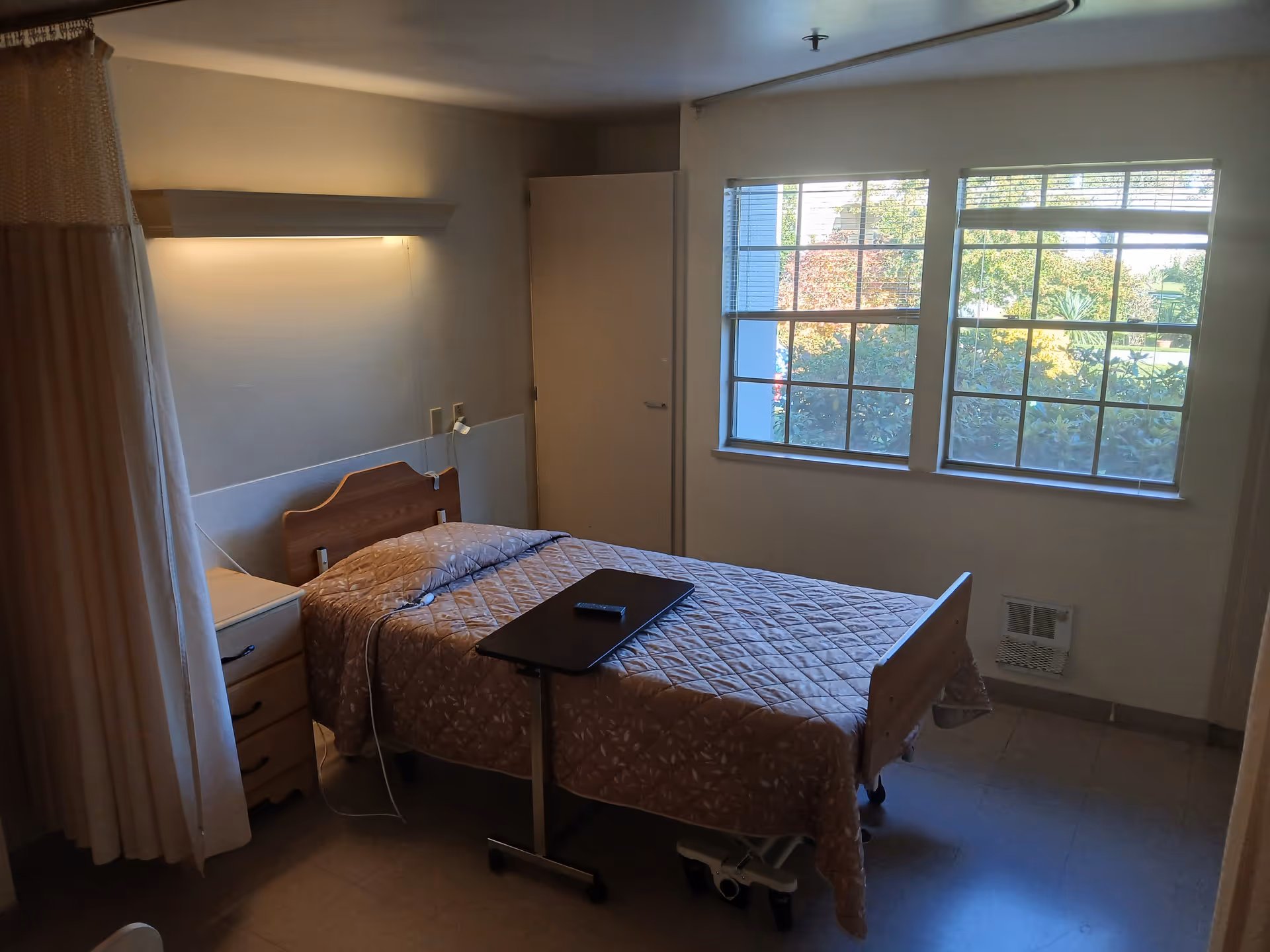 A small, simple bedroom in a healthcare facility with a single bed covered in a patterned quilt. There is a bedside table with drawers next to the bed, a curtain partially drawn on the left side, and a window on the right letting in natural light. A small overbed table with a remote control is positioned over the bed.