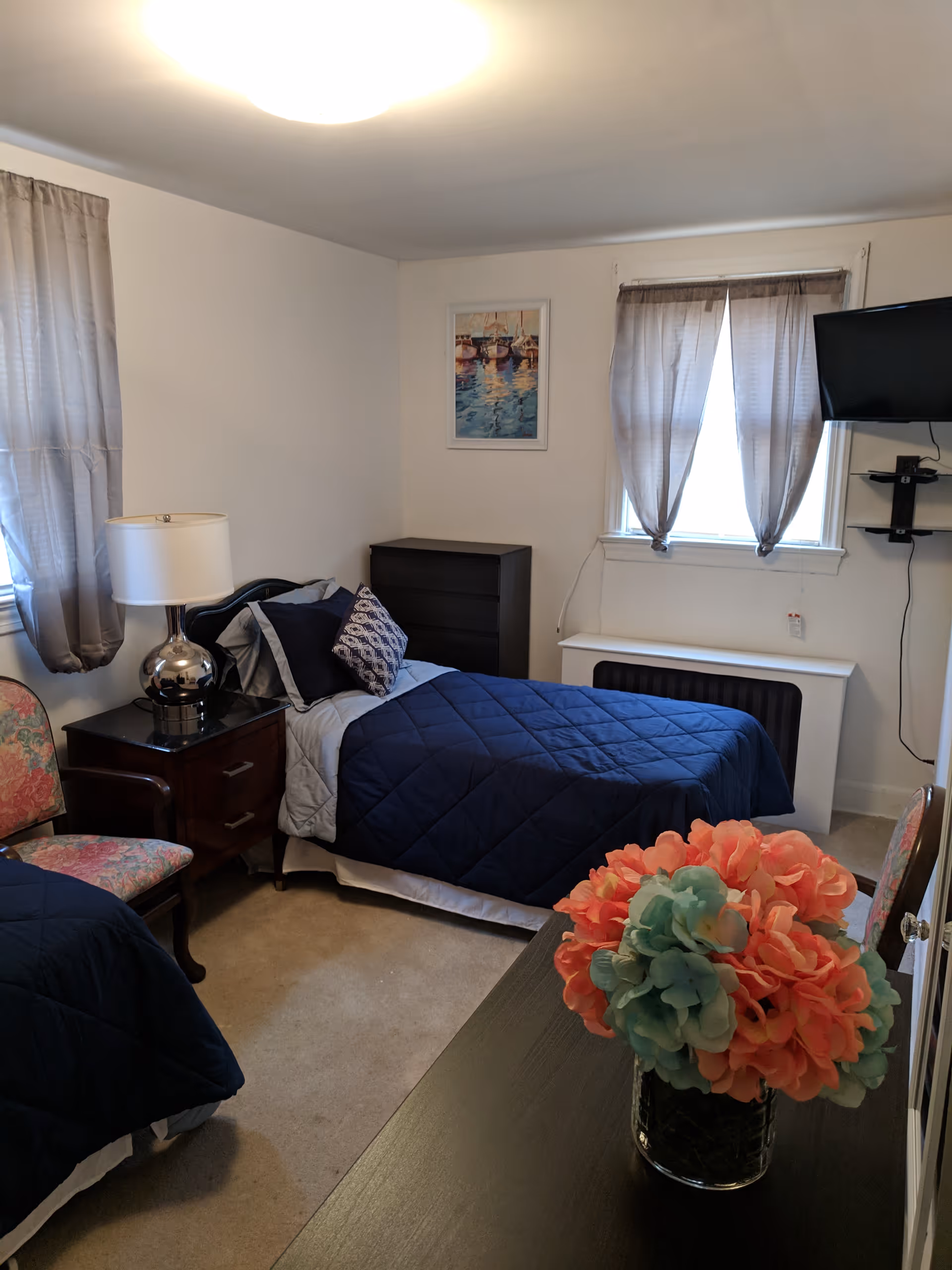 A cozy bedroom with two twin beds covered in navy blue quilts. There is a nightstand with a lamp between the beds, a floral upholstered chair, a black dresser against the wall, and a window with sheer curtains. A flat-screen TV is mounted on the wall near the window. A vase with pink and blue flowers is on a dark wooden table in the foreground.