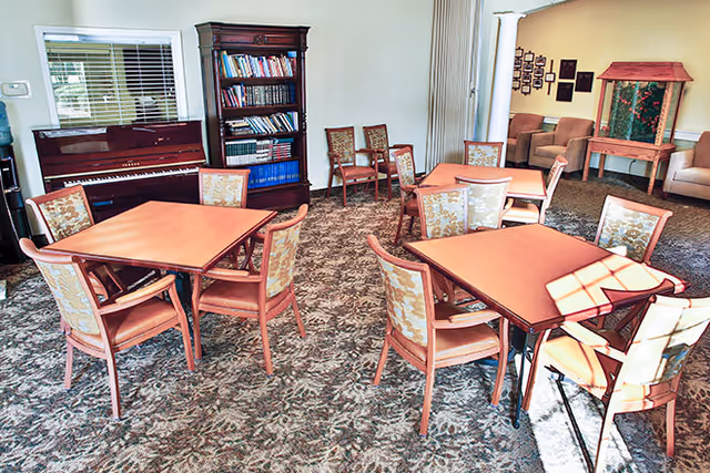 Carpeted community room with several square tables and upholstered chairs, a piano and bookshelf, and a small seating area.