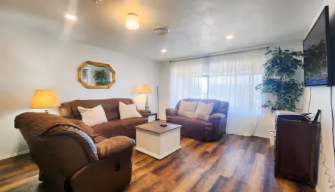 A cozy living room with two brown leather sofas and a matching armchair arranged around a white wooden coffee table. The room has wooden flooring, a large window with sheer white curtains, a floor lamp, a table lamp, a wall-mounted TV, a large potted plant, and a decorative mirror on the wall.